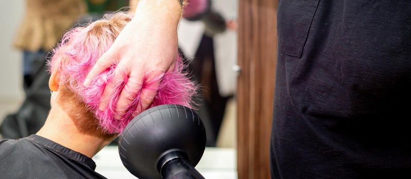 Drying short pink bob hairstyle of a young caucasian woman with a black hair dryer with the brush by hands of a male hairdresser in a hair salon, close up