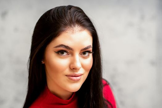 The fashionable young woman. Portrait of the beautiful female model with long hair and makeup. Beauty young caucasian woman with a black hairstyle on the background of a gray wall