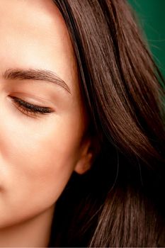 The fashionable young woman. Portrait of the beautiful female model with long hair and makeup with closed eyes. Beauty young woman with a brown curly hairstyle on the background of a green curtain