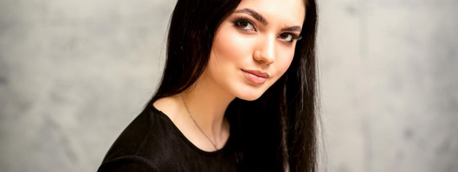 The fashionable young woman. Portrait of the beautiful female model with long hair and makeup. Beauty young caucasian woman with a black hairstyle on the background of a gray wall