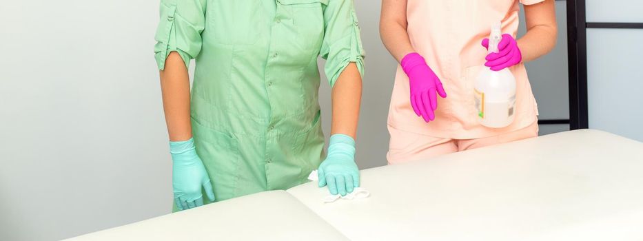 Two medical workers disinfect the patient's couch with sanitizer spray and a clean napkin. Health and hygiene concept