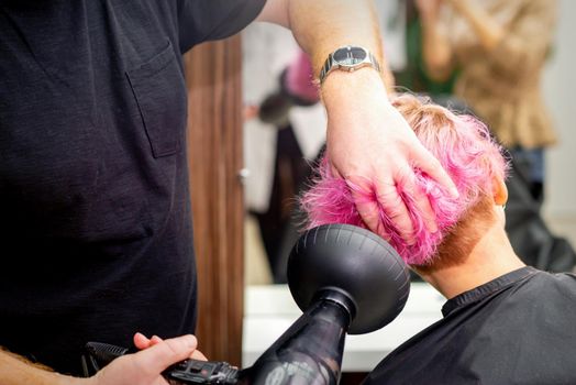 Drying short pink bob hairstyle of a young caucasian woman with a black hair dryer with the brush by hands of a male hairdresser in a hair salon, close up