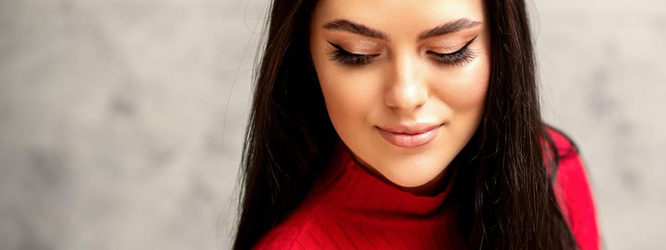 The fashionable young woman. Portrait of the beautiful female model with long hair and makeup with eyelash extensions. Beauty young woman with a black hairstyle on the background of a gray wall