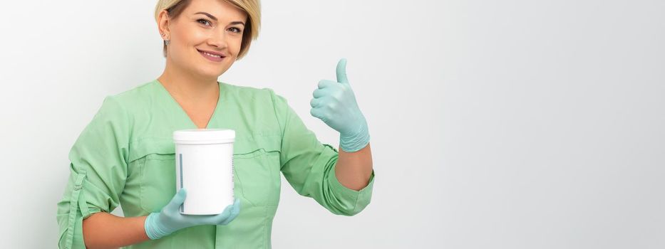 Beautician in gloves holding a jar of the cosmetic product showing thumb up on a white background