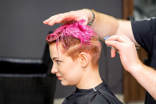 Haircut of dyed short pink wet hair of young caucasian woman by a male hairdresser in a barbershop