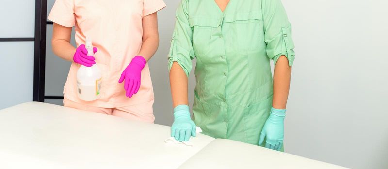 Two medical workers disinfect the patient's couch with sanitizer spray and a clean napkin. Health and hygiene concept