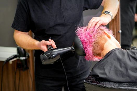 Drying short pink bob hairstyle of a young caucasian woman with a black hair dryer with the brush by hands of a male hairdresser in a hair salon, close up