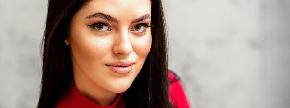 The fashionable young woman. Portrait of the beautiful female model with long hair and makeup. Beauty young caucasian woman with a black hairstyle on the background of a gray wall