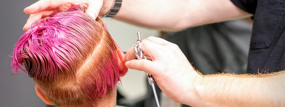 Haircut of dyed short pink wet hair of young caucasian woman by a male hairdresser in a barbershop