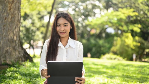 Asian woman sitting on bench in the park with computer tablet and smiling to camera.