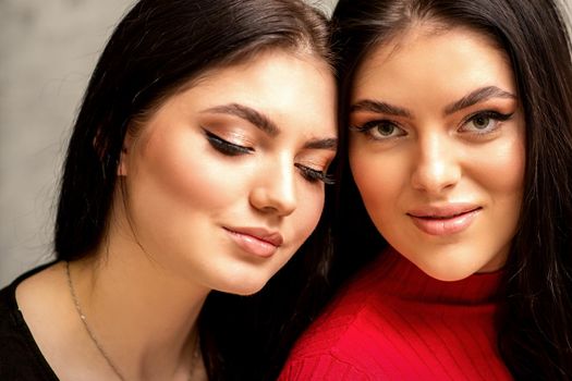 Two fashionable young women. Portrait of the two beautiful female models with long hair and makeup. Two beauty young caucasian women with a black hairstyle on the background of a gray wall