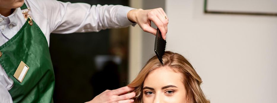 Hairdresser combing long hair of young caucasian woman looking at the camera and smiling in beauty salon