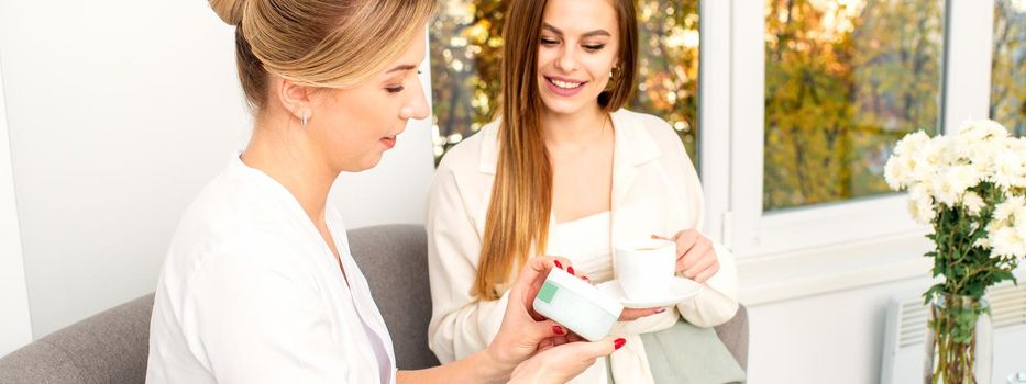 Beautician offering product for the young woman holding a white plastic jar with a cream sitting on the sofa