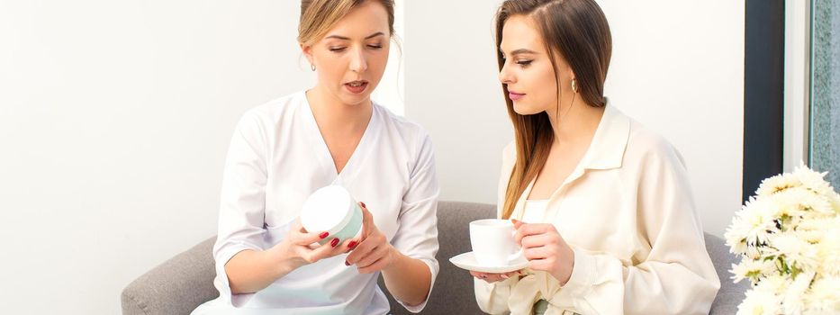 Beautician offering product for the young woman holding a white plastic jar with a cream sitting on the sofa