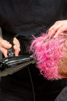 Drying short pink bob hairstyle of a young caucasian woman with a black hair dryer with the brush by hands of a male hairdresser in a hair salon, close up