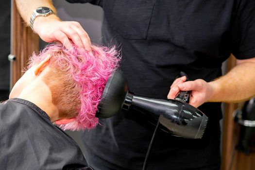 Drying short pink bob hairstyle of a young caucasian woman with a black hair dryer with the brush by hands of a male hairdresser in a hair salon, close up