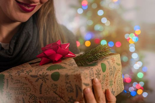 A joyful woman holds a decorative gift in her hands. Close up.Blurred Christmas tree on background. Christmas card with space for text .