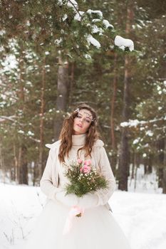 Beautiful bride in a white dress with a bouquet in a snow-covered winter forest. Portrait of the bride in nature.