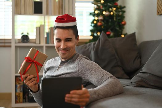 Man wearing Santa hat showing Christmas gift box and having video call with family or his friend on digital tablet.