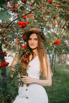 A beautiful blonde girl in a hat with long hair, on a walk in the autumn season. Portrait of a woman at a rowan tree.
