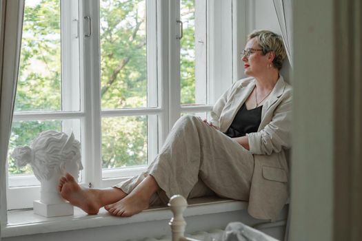 A middle-aged woman in a beige suit and black tank top sits mysteriously and looks out the window on the windowsill. Green trees outside.