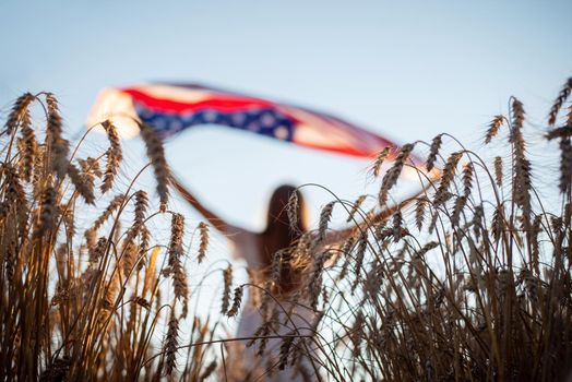 Young patriotic girl holding flag of USA in the field of rye. Focus on rye in the foreground