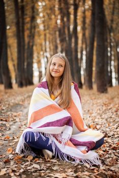 Portrait of young girl sitting on golden leaves in the autumn park and looking to the camera