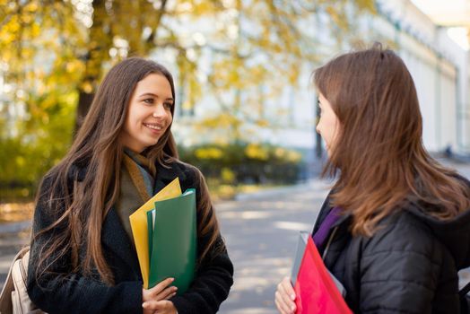 Two cheerful female students having conversation in university campus in sunny Autumn morning