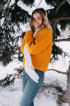 Teen blonde in a yellow sweater outside in winter. A teenage girl on a walk in winter clothes in a snowy forest.