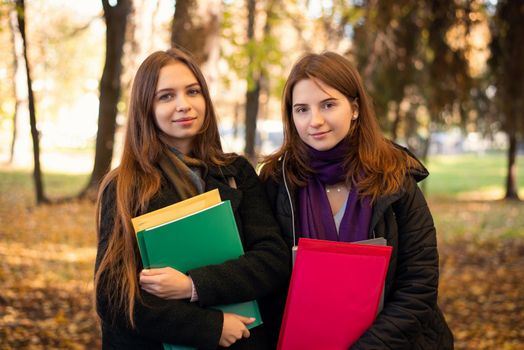 Portrait of two students with books, folders standing in the Autumn park smiling to the camera