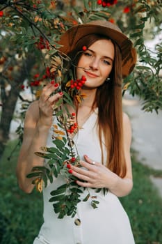 A beautiful blonde girl in a hat with long hair, on a walk in the autumn season. Portrait of a woman at a rowan tree.
