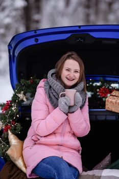 A woman in winter clothes drinks a hot drink sitting in a car decorated in a New Year's way. Traveling by car through the forest. A trip before Christmas.