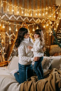 A little girl with her mother in a cozy home environment on the sofa next to the Christmas tree. The theme of New Year holidays and festive interior with garlands and light bulbs