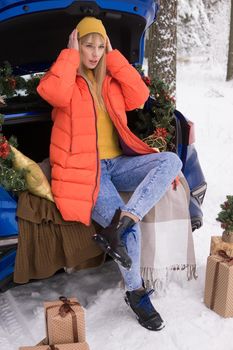 A woman in a winter snow-covered forest in the trunk of a blue car decorated with Christmas decor. The concept of Christmas and winter holidays.
