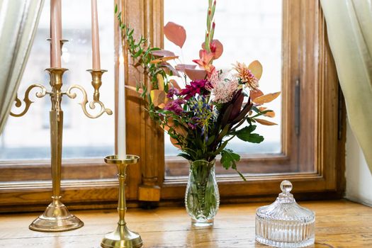 Two burning candles in a bronze candlestick an open old book and a bouquet of colorful flowers on the windowsill next to the window in the evening, close up , indoors.