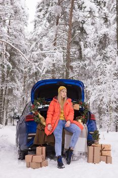 A woman in a winter snow-covered forest in the trunk of a blue car decorated with Christmas decor. The concept of Christmas and winter holidays.