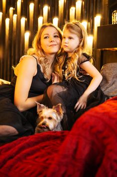 Mom and daughter in festive attire on the couch with their dog next to a Christmas tree decorated with garlands, balloons and Christmas toys. The concept of winter holidays is Christmas and New Year holidays. Magical festive atmosphere.