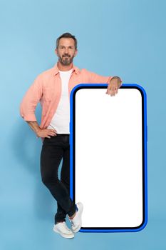 Middle aged fit man leaned on huge, big smartphone with white screen in blue case happy smiling on camera wearing peach shirt and black jeans isolated on blue background. Mobile app advertisement.