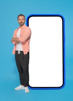 Posing with arms crossed in front big smartphone with white screen in blue case middle aged grey haired man wearing peach shirt and jogger pants isolated on blue background. Mobile app advertising.