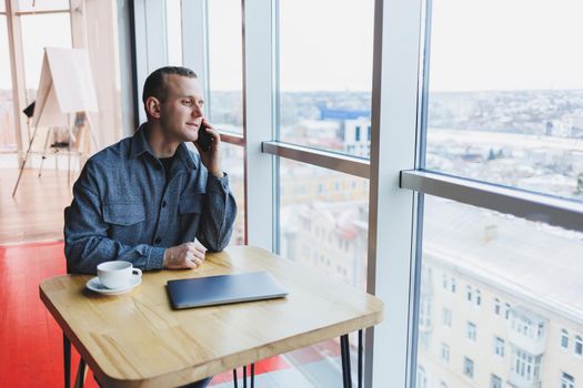 Happy man with coffee calling via smartphone while remote working laptop at work desk, cheerful freelancer enjoying coffee break while working remotely