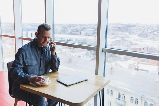 Happy man with coffee calling via smartphone while remote working laptop at work desk, cheerful freelancer enjoying coffee break while working remotely