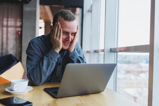 A young handsome man sits at a table and looks with surprise at the screen of his laptop, remote work