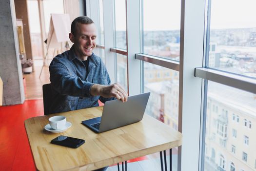 Successful happy business man is sitting at a table in a cafe, holding a cup of coffee and using a laptop.