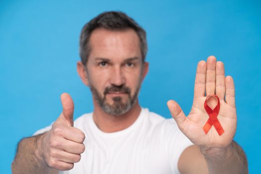 Thumb up gesture mature man with red ribbon bow AIDS awareness symbol on a palm wearing a white t-shirt isolated on a blue background. Modern medicine and healthcare. AIDS awareness concept.
