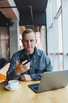 Smiling male entrepreneur in casual wear sitting at table with laptop and talking on smartphone in modern workspace with large window during day