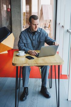 A young successful businessman in a suit drinks coffee and works in a cafe on wireless free internet on a laptop, sitting alone at a table in a cafe.