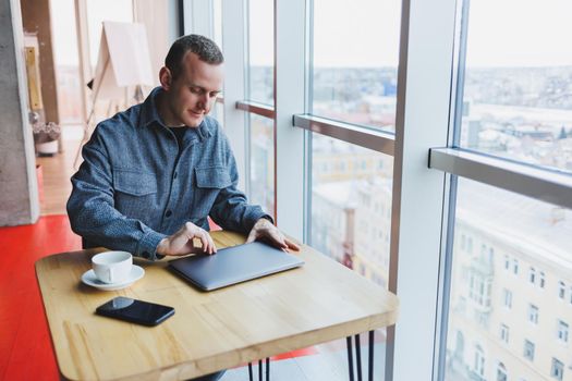 A young successful businessman in a suit drinks coffee and works in a cafe on wireless free internet on a laptop, sitting alone at a table in a cafe.
