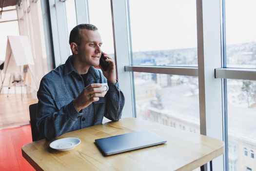 Happy man with coffee calling via smartphone while remote working laptop at work desk, cheerful freelancer enjoying coffee break while working remotely