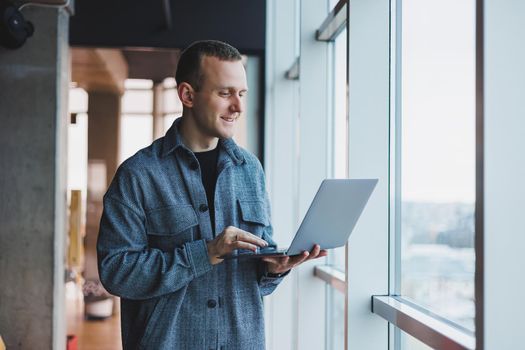Portrait of a smart intelligent male manager, he is holding a laptop for research in the office, a business man in elegant clothes playing on the touchpad. Standing in the office by a large window