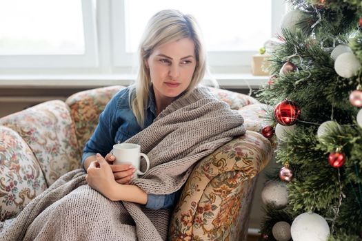 Young hispanic girl drinking coffee sitting on the sofa by christmas tree at home